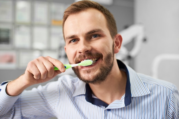 Young attractive male is smiling to the camera while cleaning his teeth with a tooth brush in a modern dental office.