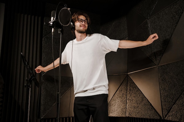 Young attractive male singer in headphones dancing during record new song in modern sound studio. Young musician singing in microphone