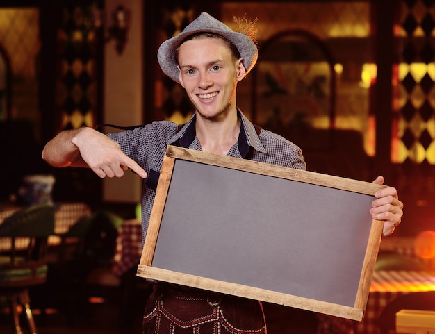 A young attractive male waiter in Bavarian clothes and wearing a hat with a feather holding a chalkboard 