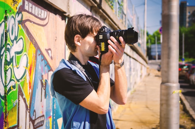 Young attractive man in a black T-shirt and blue vest takes pictures on a city street