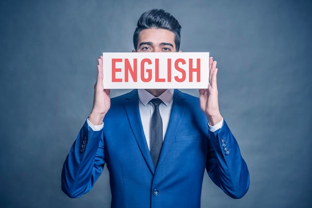 Photo young attractive man in a blue suit holding a white sign with red inscription english on a gray background toned