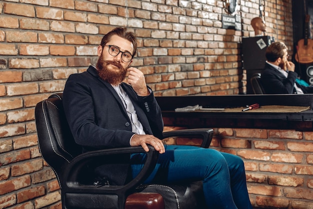 Young and attractive man in a jacket, glasses, with  sitting on a leather armchair and looking away.