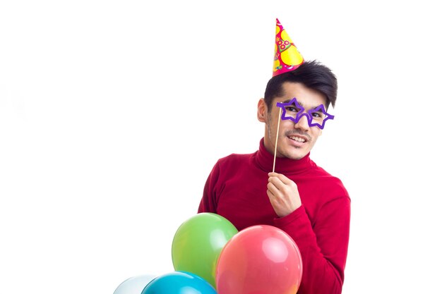 Young attractive man in red shirt with celebrating hat holding many colored balloons in studio