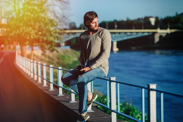 Young attractive man sitting on railing on river bank and looking away