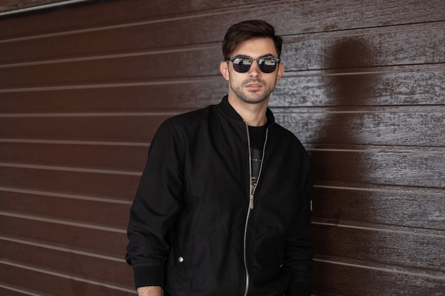 Young attractive man in stylish black jacket in trendy sunglasses with fashionable hairstyle relaxes near brown vintage wall on the street. Modern handsome guy model. Men's clothing. Style.