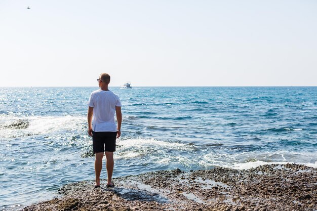 young attractive man in sunglasses in a white t-shirt and shorts stands on the shore of the mediterranean sea