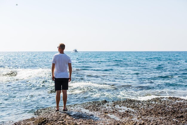 Young attractive man in sunglasses in a white t-shirt and shorts stands on the shore of the mediterranean sea