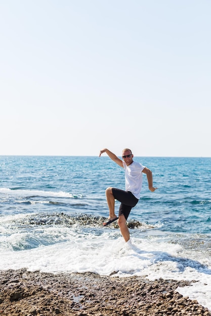 Young attractive man in sunglasses in a white t-shirt and shorts stands on the shore of the mediterranean sea