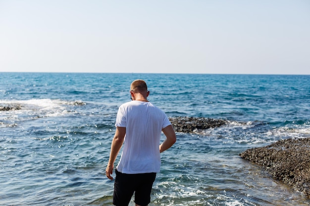 young attractive man in sunglasses in a white t-shirt and shorts stands on the shore of the mediterranean sea