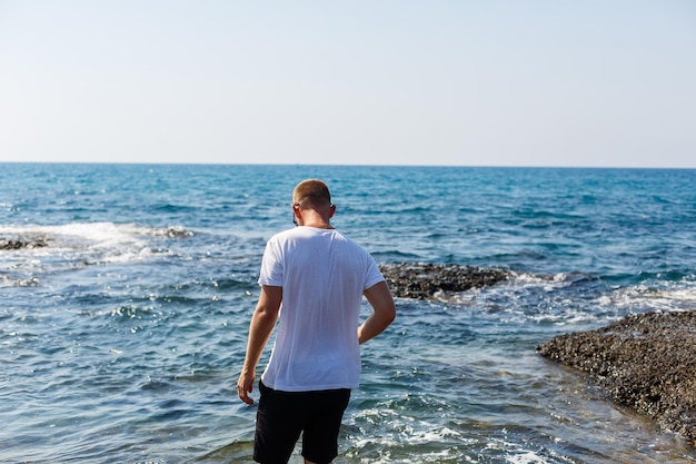 Young attractive man in sunglasses in a white t-shirt and shorts stands on the shore of the mediterranean sea