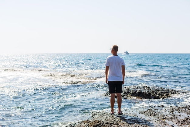 白いTシャツとショートパンツのサングラスで若い魅力的な男は、地中海の海岸に立っています
