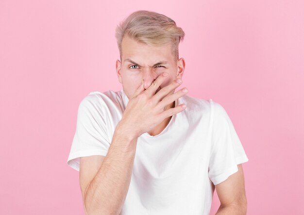 Young attractive man with disgust on his face pinches nose, something stinks, on pink