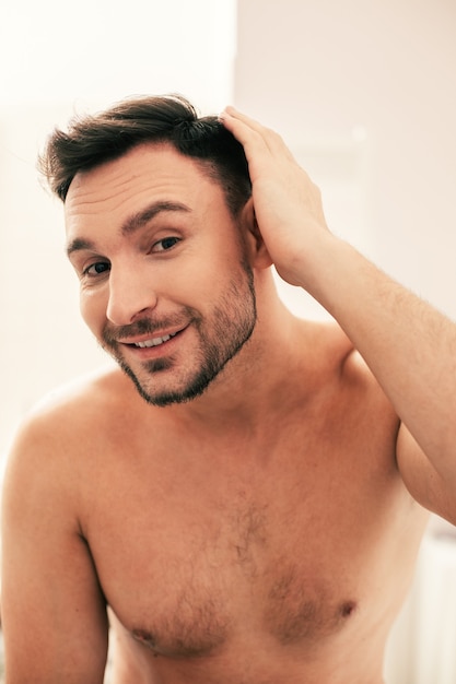 Young attractive man without a T-shirt looking glad and smiling while touching his dark hair