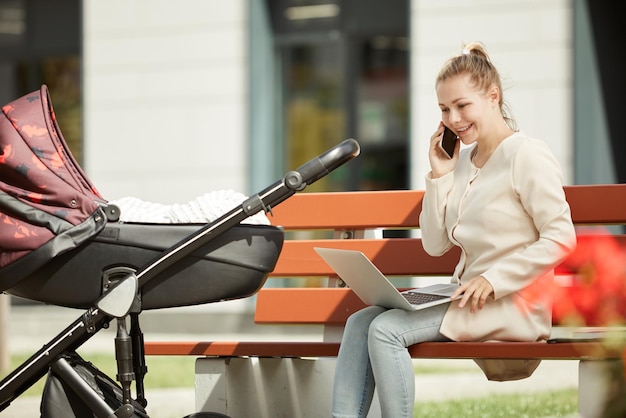 Young attractive mother working on her laptop and smartphone sitting outside on bench while baby sle