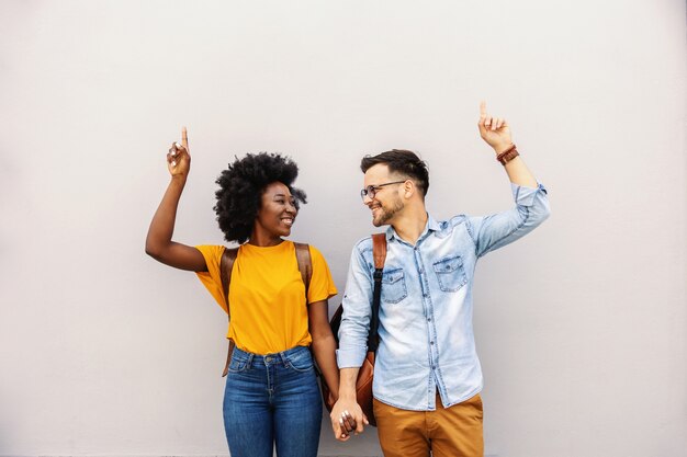 Young attractive multicultural hipster couple holding hands and pointing up.