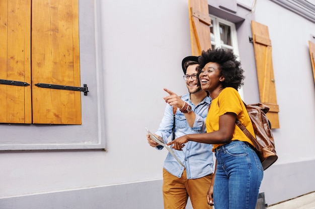 Giovani turisti multiculturali attraenti che camminano per strada e guardando splendidi edifici. donna che indica alla mappa mentre l'uomo che punta a qualcosa.