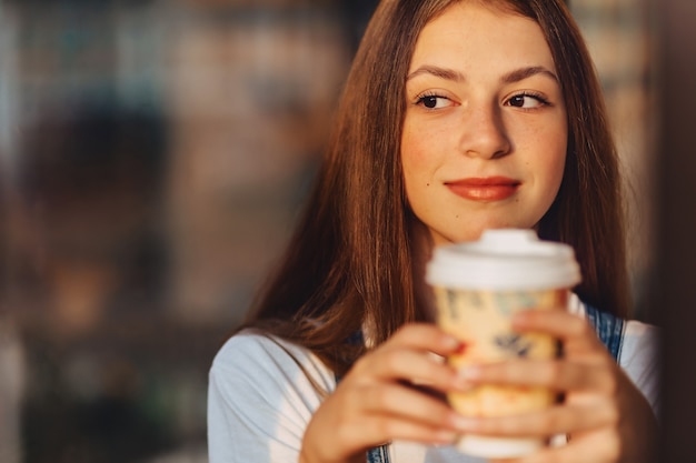 Молодая привлекательная милая девушка в кафе с кофе отдыхает на утреннем стекле