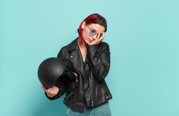 Фото Молодая привлекательная женщина с рыжими волосами чувствует скуку, разочарование и сонливость после утомительной, скучной и утомительной работы, держа лицо рукой. концепция мотоциклистов