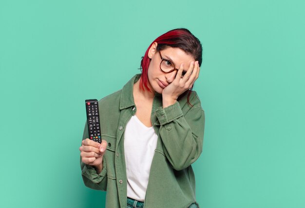 Giovane donna attraente dai capelli rossi che si sente annoiata, frustrata e assonnata dopo un compito noioso, noioso e noioso, tenendo il viso con la mano e tenendo in mano un telecomando della tv