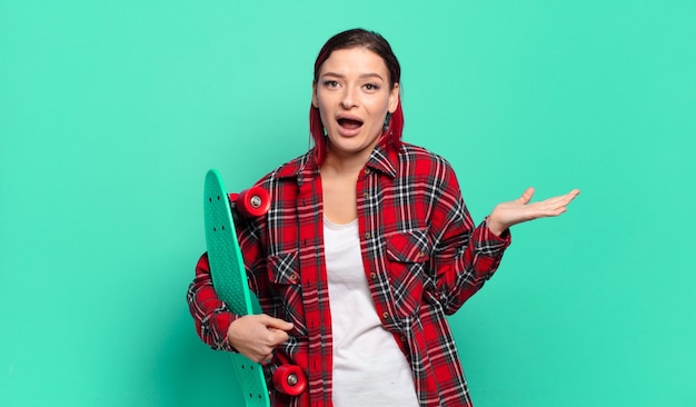 Young attractive red hair woman feeling happy, excited, surprised or shocked, smiling and astonished at something unbelievable and holding a skate board