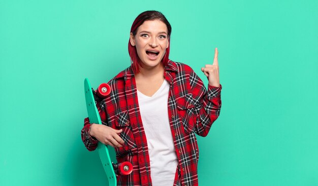 Young attractive red hair woman feeling like a happy and excited genius after realizing an idea, cheerfully raising finger, eureka! and holding a skate board