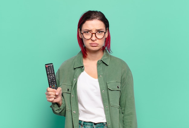 Young attractive red hair woman feeling sad and whiney with an unhappy look, crying with a negative and frustrated attitude and holding a tv remote control