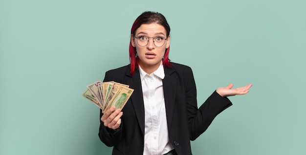 Young attractive red hair woman looking surprised and shocked, with jaw dropped holding an object with an open hand on the side. money concept