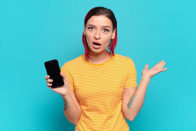 Young attractive red hair woman looking surprised and shocked, with jaw dropped holding an object with an open hand on the side and showing her cell