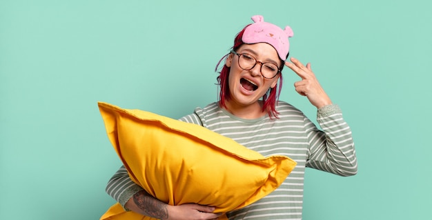 Young attractive red hair woman looking unhappy and stressed, suicide gesture making gun sign with hand, pointing to head and wearing pajamas.