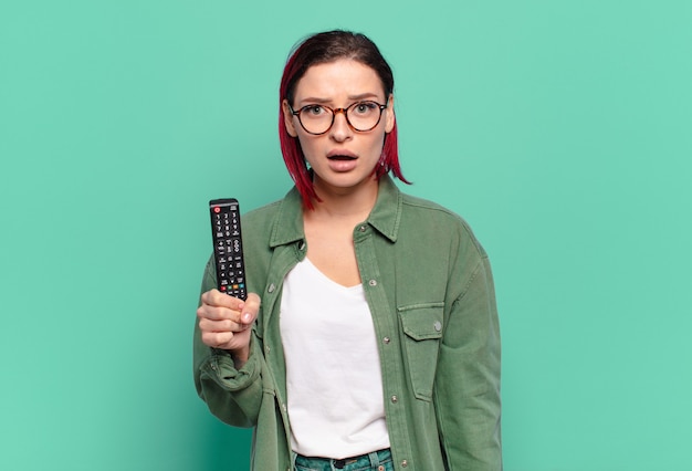 Young attractive red hair woman looking very shocked or surprised, staring with open mouth saying wow and holding a tv remote control