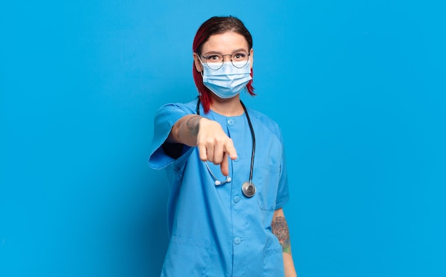 Молодая привлекательная женщина с рыжими волосами, указывая на камеру с довольной, уверенной, дружелюбной улыбкой, выбирая вас. концепция больничной медсестры