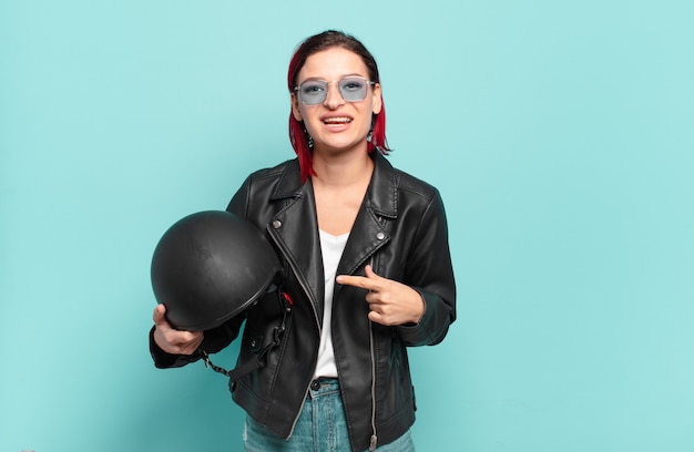 Фото Молодая привлекательная женщина с красными волосами, весело улыбаясь, чувствуя себя счастливой и указывая в сторону и вверх, показывая объект в копировальном пространстве. концепция мотоциклистов