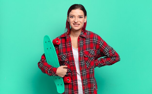 Young attractive red hair woman smiling happily with a hand on hip and confident, positive, proud and friendly attitude and holding a skate board