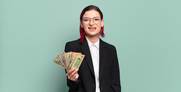 Young attractive red hair woman smiling happily with a hand on hip and confident, positive, proud and friendly attitude. money concept