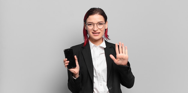 Giovane donna attraente con i capelli rossi che sorride e sembra amichevole, mostrando il numero cinque o quinto con la mano in avanti, contando alla rovescia. concetto di affari