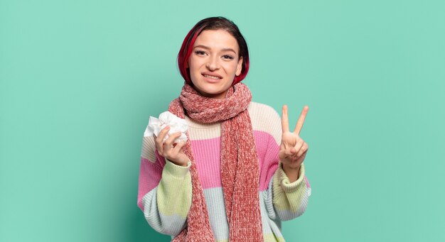 Giovane donna attraente dai capelli rossi che sorride e sembra amichevole, mostrando il numero due o il secondo con la mano in avanti, contando il concetto di influenza