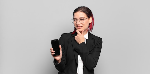 Young attractive red hair woman smiling with a happy, confident expression with hand on chin, wondering and looking to the side. business concept