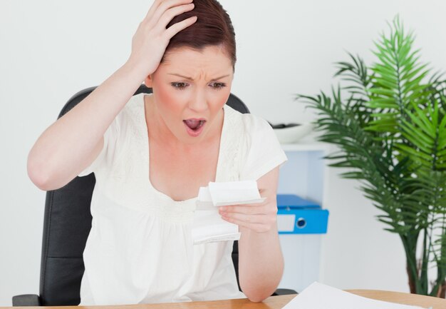 Young attractive red-haired female being surprised by the amount of the receipt