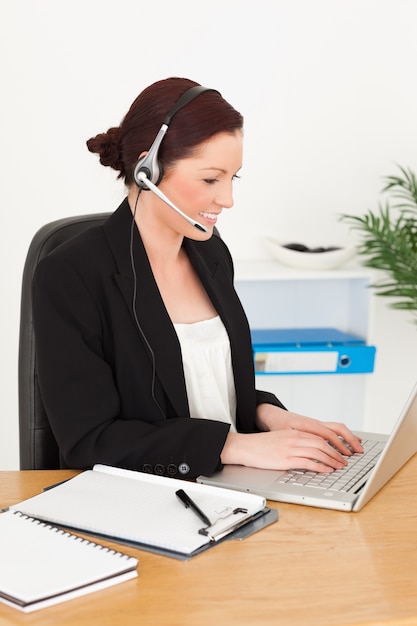 Young attractive red-haired woman in suit typing on her laptop and using headphones