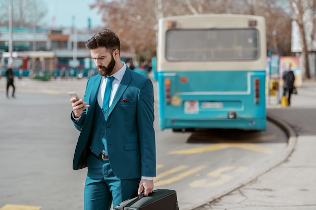 Young attractive serious businessman in suit standing on parking with luggage in hand and using smart phone while waiting on bus stop.