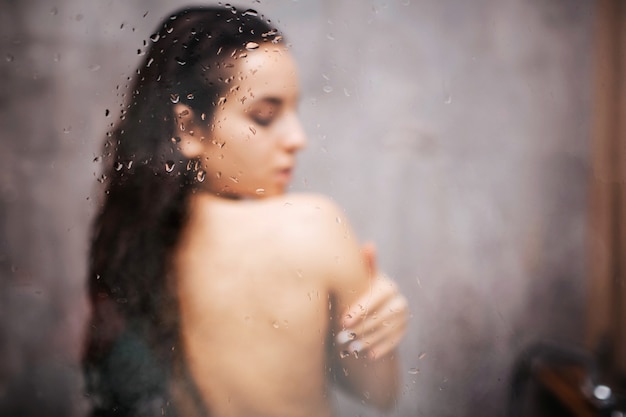 Foto giovane donna sexy attraente in doccia. in piedi di nuovo alla telecamera. abbracciala ad occhi chiusi. foto sfocata vapore acqueo sulla parete di vetro.