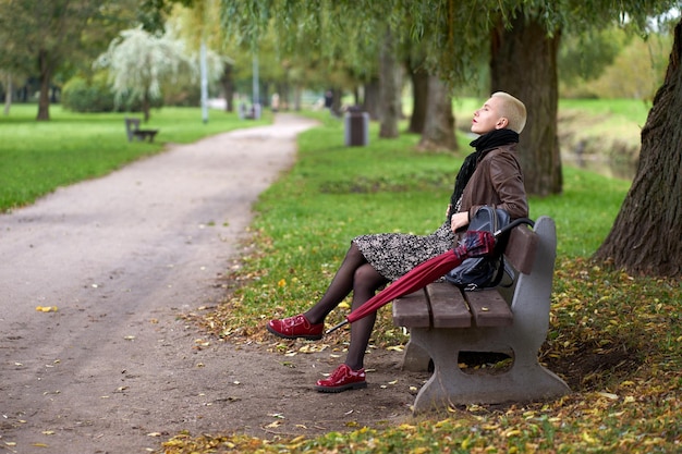 사진 빨간 우산을 들고 가을 공원의 벤치에 앉아 눈을 감고 있는 젊고 매력적인 짧은 머리 금발 여성
