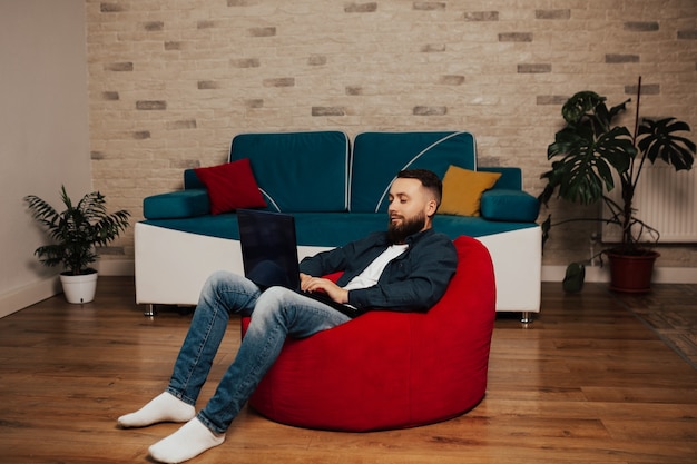 Young attractive smiling bearded guy is browsing at his laptop video, sitting at home on the cozy red armchair, wearing casual outfit.