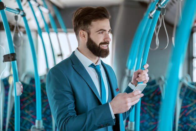 Young attractive smiling businessman in blue suit standing in public transportation and using smart phone for texting or reading message while looking trough window.