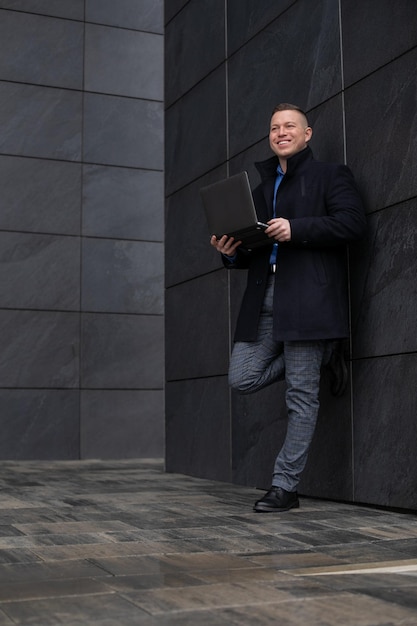 사진 검은 코트를 입은 젊은 매력적인 웃는 사업가가 무릎을 꿇고 도시 건물에 반대합니다.