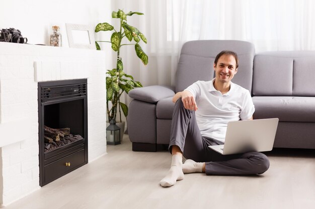 Photo young attractive smiling guy is browsing at his laptop, sitting at home on the cozy beige sofa at home, wearing casual outfit