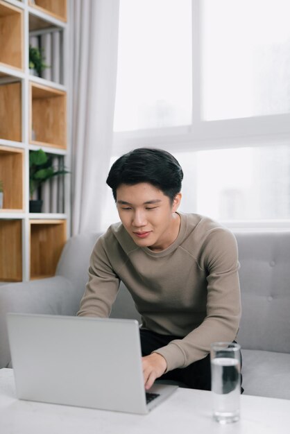 Young attractive smiling guy is browsing at his laptop sitting at home on the cozy beige sofa