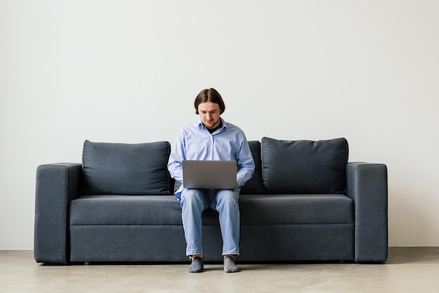 Young attractive smiling man is browsing at his laptop sitting at home on the cozy beige sofa at home wearing casual outfit