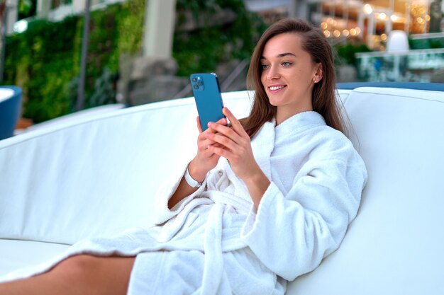 Giovane donna sorridente attraente che indossa accappatoio bianco utilizza lo smartphone per guardare video e navigare in linea mentre giaceva su un lettino durante il relax al resort termale