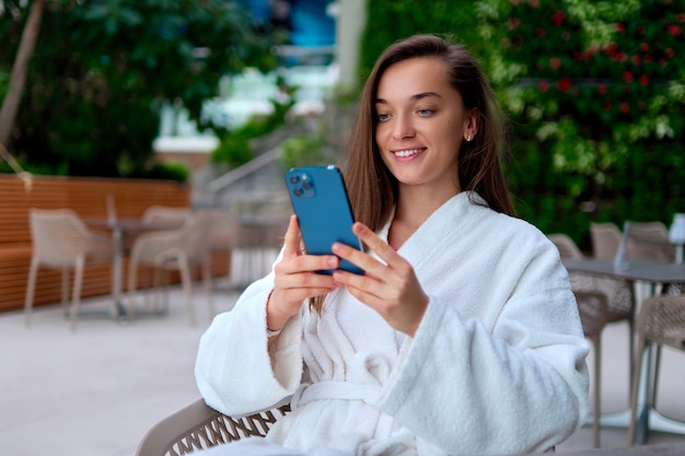 Young attractive smiling woman wearing white bathrobe using smartphone for watching video and browsing online while relaxing at spa resort